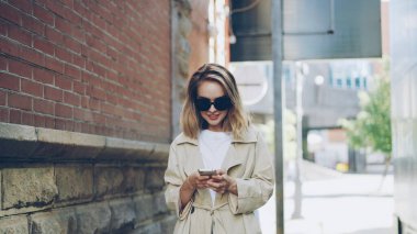 Trendy ceket ve güneş gözlük sarışın çekici genç kadınla yaşayan yürüyüş akıllı telefon kullanıyor. İnsanlar, modern teknoloji ve mutlu gençlik kavramı.
