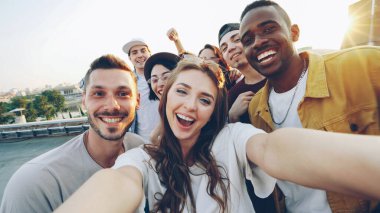 Çekici genç kadın güneş gözlüğü çatı üzerinde arkadaşlarınızla selfie alıyor, kız kamera tutuyor ve onu arkadaşları komik yapma eğleniyor iken poz yüzleri ve jestleri.