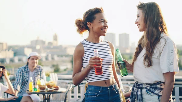 かなりアフリカ系アメリカ人の女の子は彼女の陽気な白人の友人に話しているし 屋上で野外パーティーの最中にカクテル持株ボトルを飲みます 楽しさと通信の概念 — ストック写真