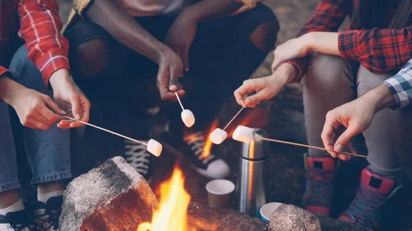 Close-up shot of burning campfire and peoples hands holding sticks with marshmallow above flame and tourists legs in sneakers getting warm near fire. Camping and food concept.