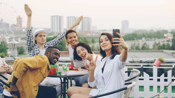かなり若い女性はスマート フォンを押し 屋上に食べ物や飲み物とテーブルに座って感情的な友人と Selfie を撮影です 美しい都市は — ストック写真