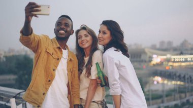 Şık Afrikalı-Amerikalı adam çatıda açık hava partisinde Sevimli beyaz kızlarla selfie alıyor, Genç adam modern smartphone kullanarak, kadın bira şişesi tutuyor.