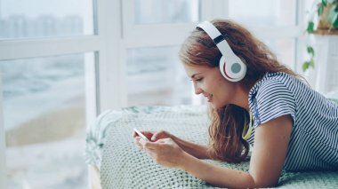 Sevimli genç kadın kulaklık müzik dinlemek ve yatağa evde istirahat smartphone ekran dokunaklı. Modern teknoloji, gençlik kültürü ve iç kavramı.