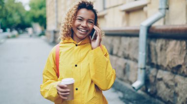 Parlak paltolu güzel Afrikalı-Amerikalı kız cep telefonu üzerinde konuşurken ve cadde boyunca şehirde yürüyüş ve zevk konuşma gülüyor take away kahve holding.