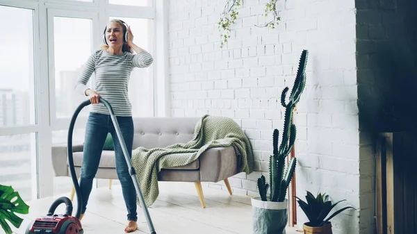 stock image Cheerful blond housewife is using vacuum cleaner during routine clean-up at home, listening to music through headphones, singing and dancing. Technology and young people concept.