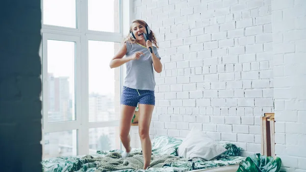 stock image Joyful young woman is dancing on bed at home wearing headphones and enjoying pop music. Double bed, brick wall, panoramic window and green plant are visible.