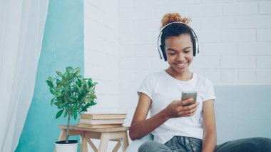 Mutlu Afrikalı-Amerikalı kadın smartphone ve evde rahat koltukta oturan gülüyor ile internet sörf, kız kulaklık aracılığıyla müzik dinlerken.