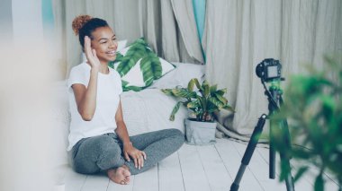 Oldukça Afrikalı Amerikalı genç vlogger sosyal medya takipçileri için video kayıt, kız kameraya tripod üzerinde konuşma ve yatak odası katta oturan el hareketi bakıyor.