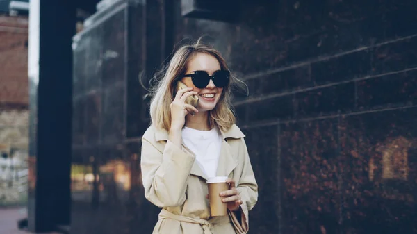 ブロンドの髪を持つ美しい若い女性が携帯電話で話しているし 近代的な都市通りに沿って歩いて 移動するドリンクや会話を楽しんで取る離れてコーヒーを飲む — ストック写真