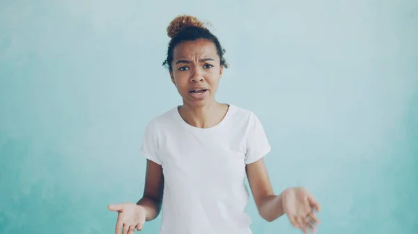 Portrait of angry African American woman talking and gesturing expressing negative emotions standing against light blue background. Feelings, reactions and people concept.