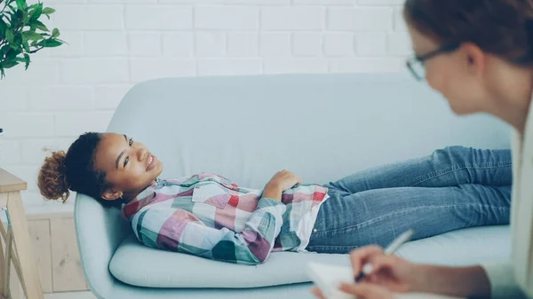 気配りのある心理学者は若いアフリカ系アメリカ人女性が 聞いて メモを作っているクライアントはソファに横になっていると笑顔で話すとの協議を保持しています — ストック写真