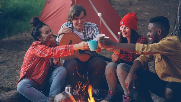 Joyful young people friends are clinking glasses with drinks sitting around fire nera tent in forest with warm marshmallow on sticks, smiling man is holding guitar.