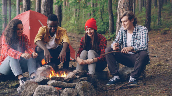 Мультиэтническая группа друзей туристы сидят вокруг огня разговаривают и смеются, молодой человек бросает дрова в огонь. Кемпинг, дружба и летняя природа
.