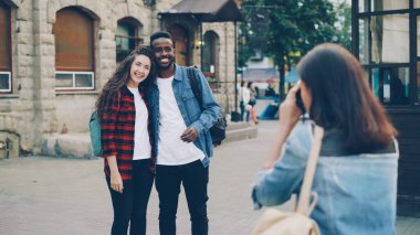 Genç bir kadın, sokakta durup kameraya poz veren güzel bir çift Afro-Amerikalı ve beyaz kız turistin fotoğraflarını çekiyor. Turizm, teknoloji ve arkadaş kavramı.