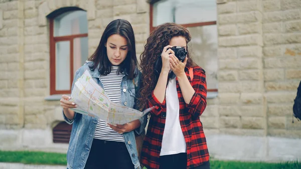 女性の観光客が写真を撮っている間 彼女の友人の魅力的な女の子は地図を見ていて 通りに立って彼女の方向を示しています 旅と写真のコンセプト — ストック写真