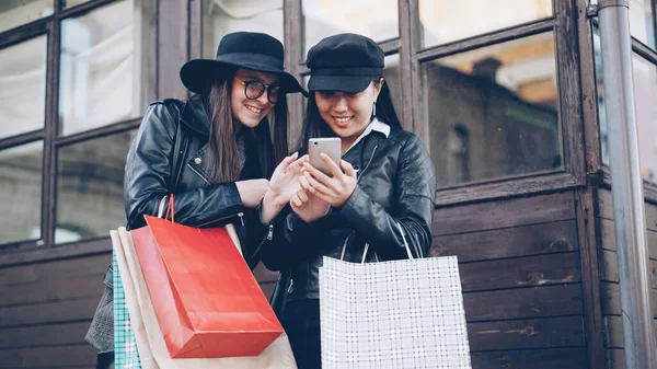 兴奋的女孩朋友们使用智能手机触摸屏站在街上 并举行购物袋在秋季的一天 现代技术与青年观念 — 图库照片