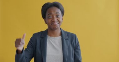 Memnun olmayan Afro-Amerikalı kadın başparmağını kaldırıyor ve kameraya bakıyor. Sarı arka planda negatif duyguları ifade ediyor.