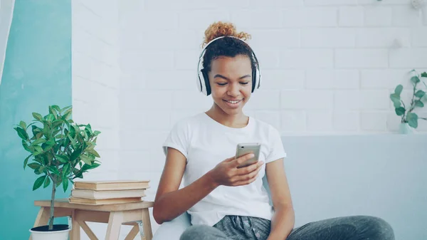 아프리카계 미국인 집에서 소파에 스마트폰으로 인터넷을 여자는 헤드폰을 음악을 듣으십시오 — 스톡 사진