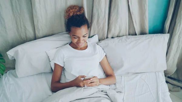 Веселая Афроамериканка Использует Смартфон Лежащий Постели Теплым Одеялом Мягкой Подушке — стоковое фото