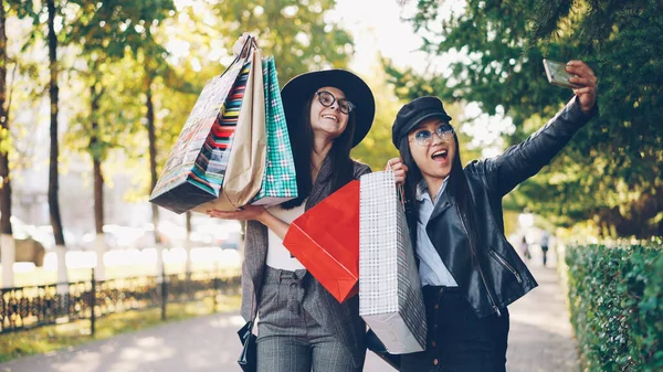 快乐的年轻女性朋友们正带着自拍的彩色纸袋站在人行道上 摆姿势 女孩们使用智能手机和穿着休闲服装 — 图库照片
