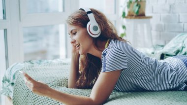 Sevimli genç kadın kulaklık müzik dinlemek ve yatağa evde istirahat smartphone ekran dokunaklı. Modern teknoloji, gençlik kültürü ve iç kavramı.