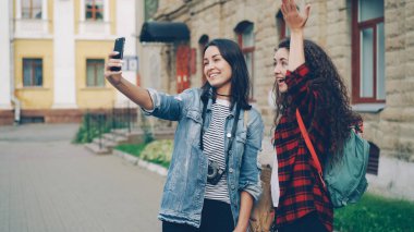 Neşeli yabancı kızlar, dışarıda duran akıllı telefonları kullanarak selfie çekiyorlar. El hareketleriyle V işaretini ve kalbi parmaklarıyla gösteriyorlar ve birlikte gülüyorlar..