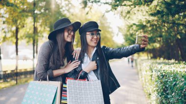 Mutlu genç kadın öğrenciler ile akıllı telefon bahsediyoruz gösteren çanta alışveriş ve ayakta sokakta sonbahar gününde gülüyor online görüntülü görüşme yapıyoruz.