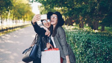 Heyecan arkadaşlar smartphone kullanarak ve alışveriş torbaları sonbahar güneşli satın almalarla holding selfie alıyor. Modern teknoloji ve gençlik yaşam tarzı kavramı.