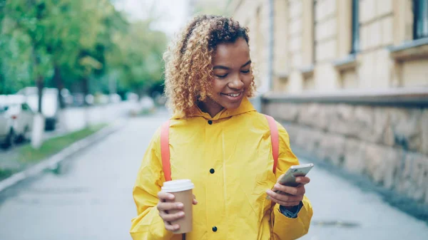 Smiling African American Girl Using Smartphone Texting Friends Holding Coffee — Stock Photo, Image
