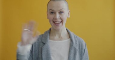 Dost canlısı genç bayan el sallıyor ve gülümsüyor sarı arka planda tek başına duran kameraya bakıyor. İnsanlar ve merhaba jest konsepti.