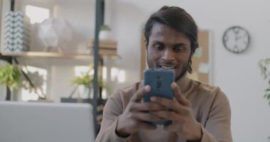 Hintli girişimcinin portresi akıllı telefon kullanarak mesajlaşarak ve gülümseyerek ofiste tek başına çalışıyor. Kablosuz teknoloji ve iş iletişimi kavramı.