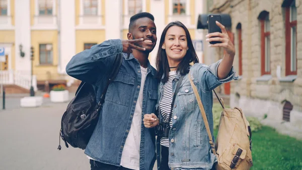 バックパックを持つ幸せな観光客の友人は 夏の日に美しい街の通りで一緒に手のジェスチャーVサインと親指の立ち上がりを示すスマートフォンを使用して自撮りしています — ストック写真