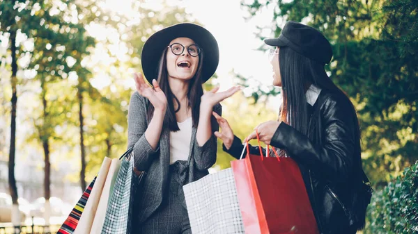 快乐的亚洲女孩与购物袋正在谈论她的女性朋友站在街上 然后显示她的购买 而高加索年轻女子表示积极 Emotons — 图库照片
