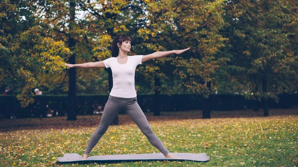 Attractive young woman is doing yoga outdoors standing on mat and practising sequence of asanas Triangle pose and Warrior position enjoying fresh air in autumn.