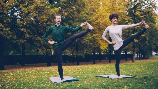 Slim young ladies are exercising outdoors in park standing on one leg practising Padangusthasana during individual class with experienced teacher. Nature and youth concept.