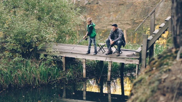 Good-looking man and his cute son are fishing in pond from wooden dock sitting on chairs with rods and talking. Loving family, common hobby, nature and generations concept.