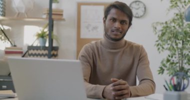 Ofiste oturmuş gülümseyen ve tek başına çalışmaktan zevk alan neşeli Hintli işadamının yavaş çekim portresi. İnsanlar ve olumlu duygu kavramı.