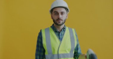 Kasklı ve yelekli bir adamın portresi. Elinde elektrikli matkap var. Sarı arka plandaki kameraya gülümsüyor. Profesyonel enstrüman ve tamirci konsepti.