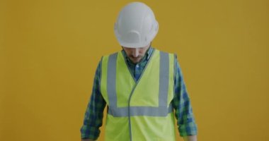 Kask ve yelek giymiş, çekiç ve penseyle el kaldıran ve sarı arka planda gülümseyerek kameraya bakan bir adamın yavaş çekim portresi.