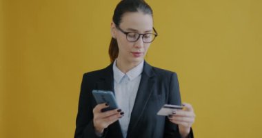 İş kadınının portresi kredi kartıyla ödemeyi sarı arka planda akıllı telefon kullanarak yapıyor. Takım elbiseli genç bir kadın, elinde plastik kartla ekrana dokunuyor ve gülümsüyor..