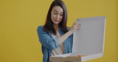 Aç genç bir bayanın portresi. Kutuyu açıyor, pizzaya bakıyor ve sarı arka planda gülümseyen dilimler yiyor. Fast food ve teslimat konsepti.