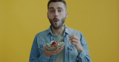 Sebze salatası yiyen neşeli genç adamın portresi sarı arka planda gülümseyen sağlıklı ve lezzetli yiyeceklerin tadını çıkarıyor. Beslenme ve vejetaryen konsepti.