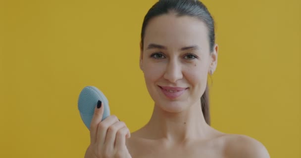 黄色の背景にカメラを見て笑顔化粧品デバイスとかわいい若い女性のマッサージ顔のスローモーション肖像画 現代のテクノロジーとスキンケアの概念 — ストック動画