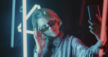 Akıllı telefon kamerası için poz veren genç bir kadının neon ışıklı alanda selfie çekmesi. Modern teknoloji ve oto portre konsepti.