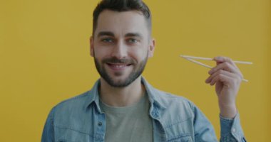 Elinde tahta çubuklar tutan gülümseyen adamın portresi sarı arka planda yemek için bekliyor. Doğu mutfağı ve beslenme konsepti.