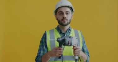 Kasklı, elektrikli matkap tutan inşaat işçisinin portresi sarı arka planda enstrümanla hareket ediyor. Ekipman ve enstrüman kavramı.