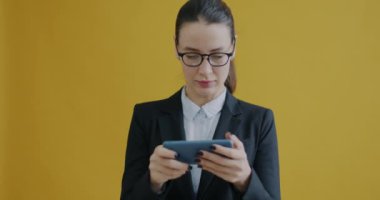 Sarı arka planda akıllı telefonlu video oyunu oynayan kaygısız bir iş kadınının yavaş çekim portresi. Modern teknoloji ve eğlence etkinliği kavramı.