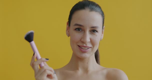 玩世不恭的女人拿着化妆刷触摸鼻子 微笑着在黄色背景下看着相机的慢动作肖像画 — 图库视频影像