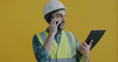 Üniformalı müteahhit cep telefonuyla konuşuyor ve sarı arka plandaki proje belgelerine bakıyor. İnşaat işi ve iş konsepti.