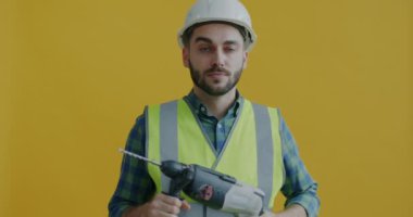 Güvenlik üniformalı, elinde elektrikli matkapla dolaşan ve sarı arka planda eğlenen silah gibi ateş eden oyuncu portresi. Alet ve tamirci kavramı.